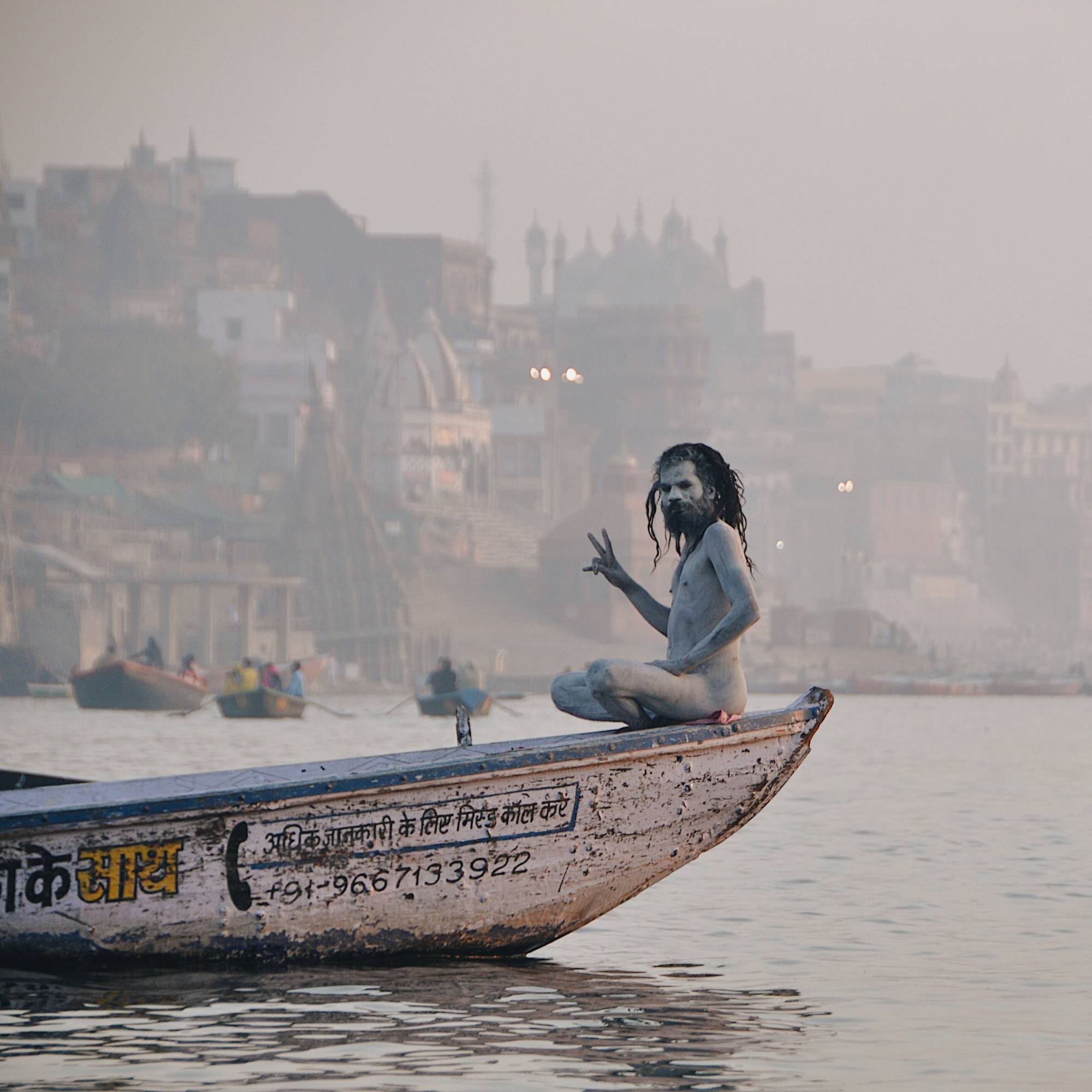 Colored photograph of man painted white, giving a "peace sign" sitting on the very tip of the bow of a long boat or canoe. Foggy city and other boats behind him,